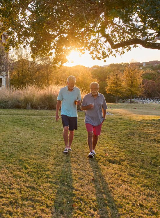 two casually dressed senior men carrying cocktails walk in the grass at sunset at Querencia Senior Living Community