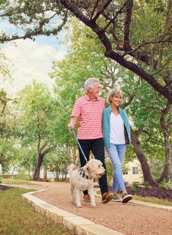 smiling senior couple with arms around one another walk their small white dog along a tree-lined path at Querencia Senior Living Community