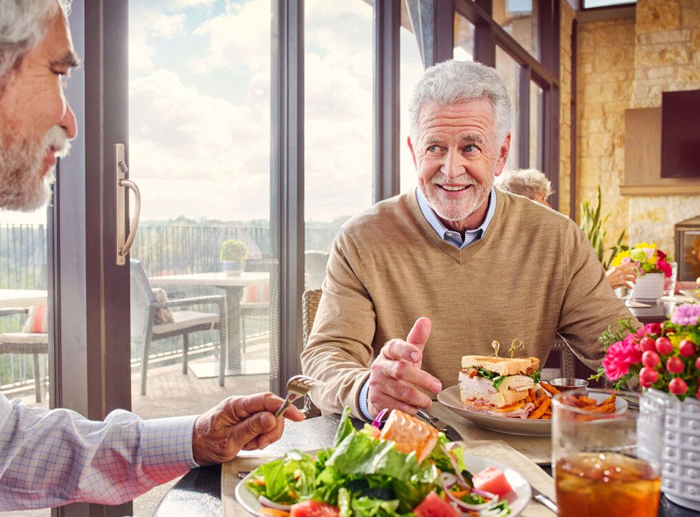 two male seniors converse while enjoying a lavish dinner with friends in the dining room at Querencia Senior Living Community