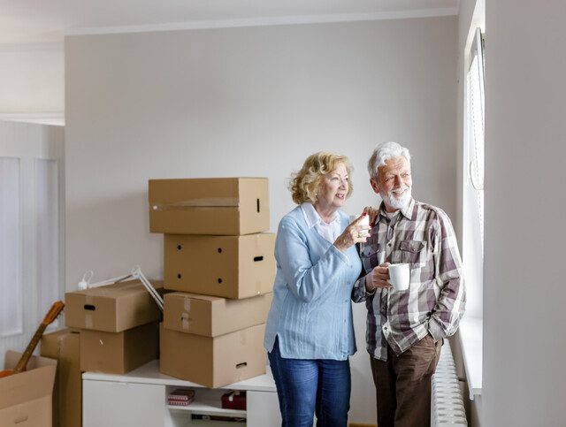 smiling senior couple holding coffee and enjoying the view through their window, surrounded by packed moving boxes