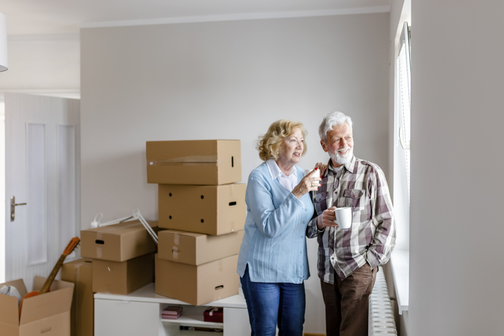 smiling senior couple holding coffee and enjoying the view through their window, surrounded by packed moving boxes