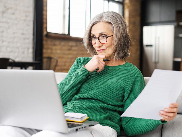 senior woman in green sweater sitting on her couch reviewing information on her laptop
